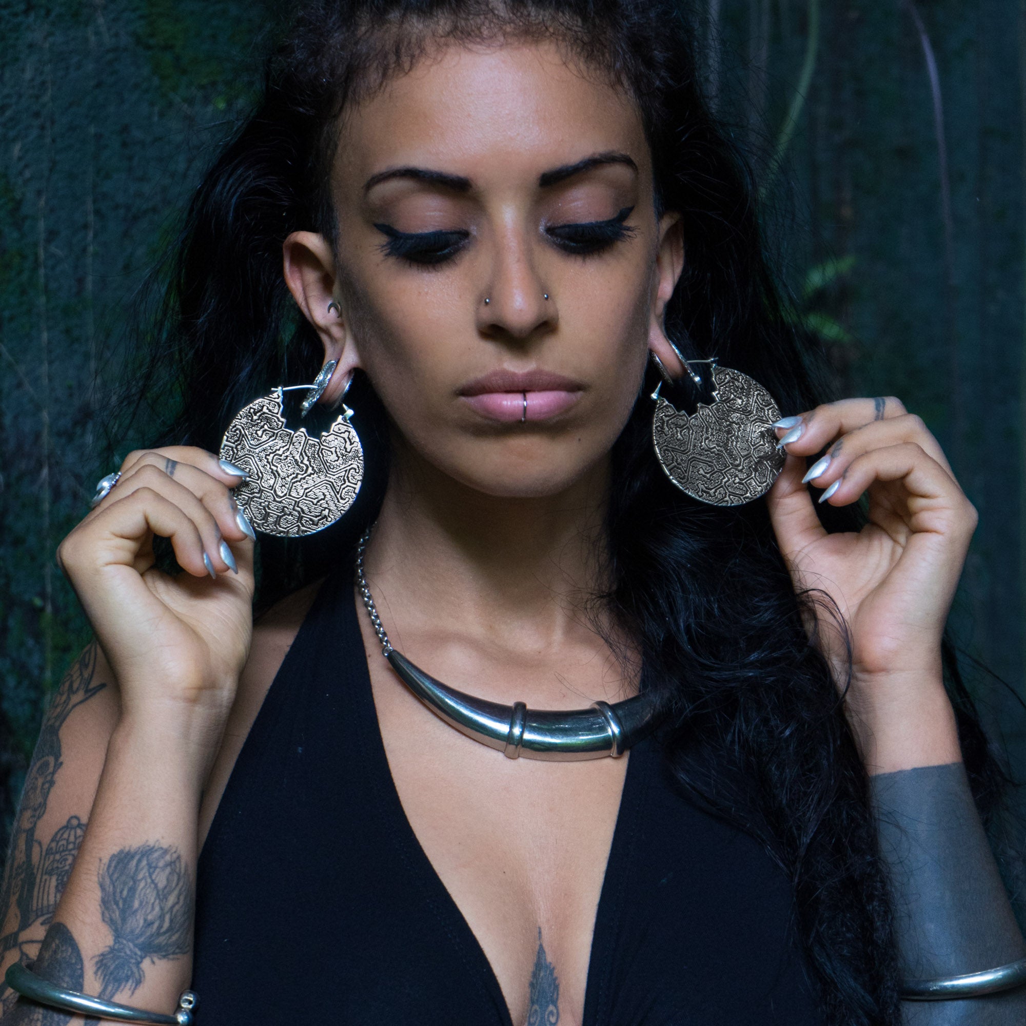 A woman with long dark hair, piercings, and tattoos is wearing a black top and a large metal necklace. She holds up two PANCHA Oversized Tribal Shipibo Disc Earrings in Silver by Kaya Movement. With her eyes closed, she stands against a dark, textured background.